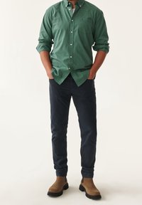 Camisa de cuadros verdes con cuello y botones, pantalones navy y botas de tobillo marrones. La camisa tiene cuello y cierre frontal de botones. Textura de tela suave.