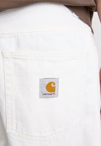 Hvite denimshorts med en baklomme med en grå etikett. Teksten på etiketten viser et gult logo og "carhartt" i mørk tekst.