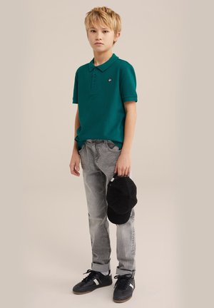 Türkises Polohemd mit kleinem Logo, graue Jeans und schwarze Cordmütze in der Hand. Schwarze Sneakers mit weißen Akzenten. Kurze Haare.