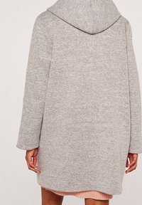 Szary płaszcz z kapturem o teksturowanym wykończeniu, luźnym kroju i długich rękawach. Charakterystyczne przeszycia wzdłuż szwów oraz zaokrąglony dół.