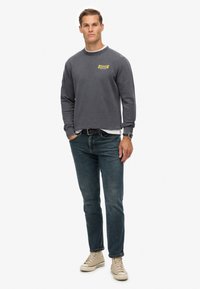 Hombre de pie con sudadera gris con logo amarillo, jeans azules, zapatillas altas beige, cinturón negro y reloj en la muñeca, con las manos en los bolsillos.