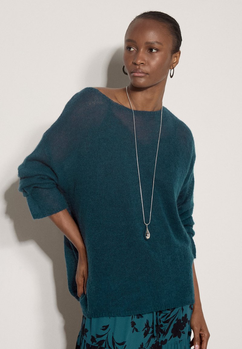 Femme portant un pull ample couleur sarcelle, un long collier en argent et une jupe bleue fleurie, debout avec une main dans la poche contre un mur clair.