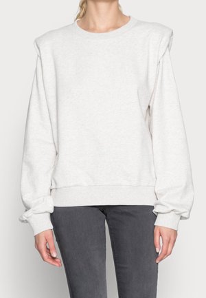 Kobieta ubrana w jasnoszary sweter z wyściełanymi ramionami i ciemnoszare dżinsy, stojąca z ramionami swobodnie opuszczonymi wzdłuż ciała.