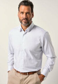 Muž s vousy, oblečený v bledě pruhované košili zasunuté do béžových kalhot s hnědým opaskem a hodinkami, stojící s rukama v kapsách.