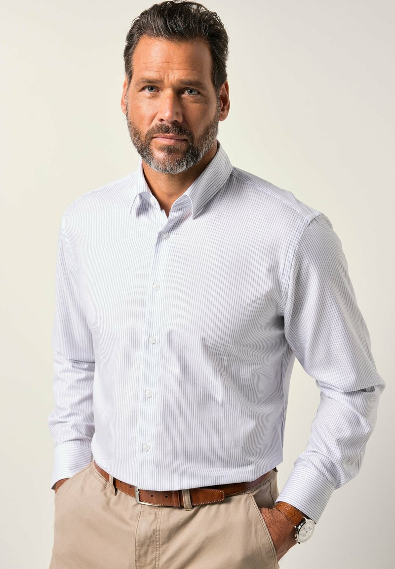 Muž s vousy, oblečený v bledě pruhované košili zasunuté do béžových kalhot s hnědým opaskem a hodinkami, stojící s rukama v kapsách.