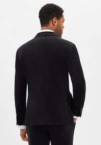 Blazer nero realizzato in tessuto liscio, con vestibilità sartoriale, spacco singolo sul retro e polsini con bottoni che lasciano intravedere i polsini della camicia bianca.
