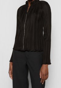 Giacca nera con zip, caratterizzata da pieghe verticali, colletto alto e orlo arricciato. Presenta una zip argentata e una trama sottile. Indossata con pantaloni neri.