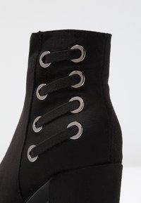 Botín de ante negro con tacón cuadrado, con seis ojetes metálicos, cordones a juego y una textura suave.