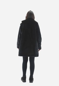 Sort faux fur frakke med bred krave, lange ærmer og en afslappet pasform, båret over sorte leggings og ankelstøvler.