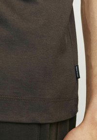 T-shirt in cotone marrone scuro con una texture liscia, orlo dritto e un piccolo etichetta cucita nella cucitura laterale. Nessun motivo o grafica.
