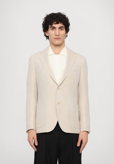 Beige tailored blazer with two buttons, notch lapels, and front pockets. Worn over a light-colored shirt, paired with dark trousers.