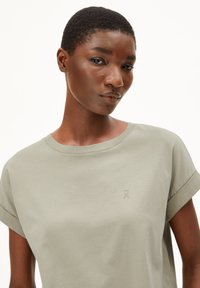 T-shirt vert olive clair en coton doux. Présente des manches retroussées et un logo brodé discret. Texture lisse et coupe décontractée.
