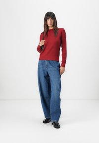 Suéter de punto rojo acanalado con mangas largas, combinado con unos jeans de denim azul sueltos. Mocasines negros completan el conjunto. Fondo sencillo.