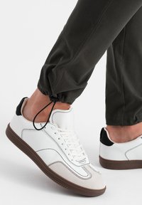 Witte sneakers met een glad leren bovenwerk, een lichtgrijze suède neuskap, een bruine rubberen zool en zwarte accenten op de hiel.