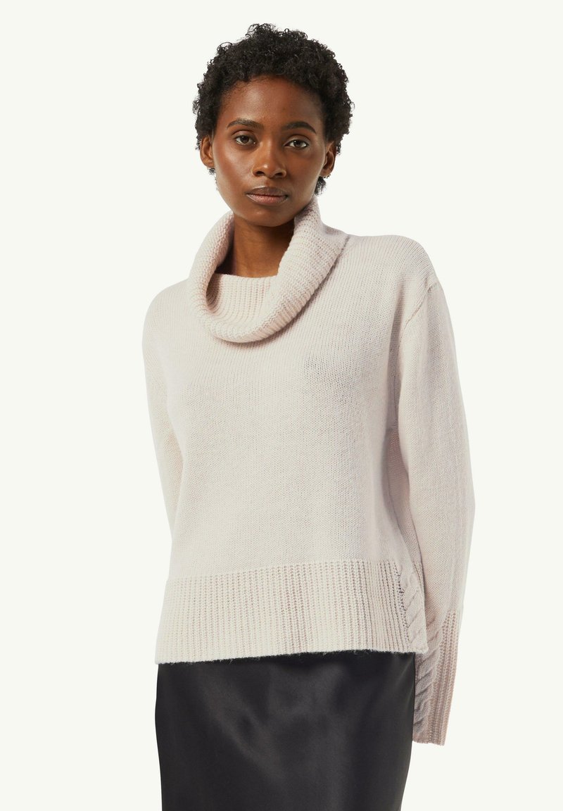 Jasnoróżowy dzianinowy sweter z dekoltem typu cowl, prążkowanym wykończeniem i bocznymi rozcięciami. Miękka struktura i luźny krój, zestawiony z czarną satynową spódnicą.