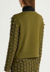 Pull vert olive avec détail texturé et superposé sur les manches, souligné de contours violets ; coupe ajustée avec un col noir au niveau de l'encolure.