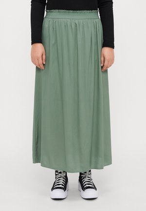 Personne portant une jupe longue taille haute vert sauge, un top noir à manches longues et des baskets montantes noires et blanches, debout sur un fond blanc.