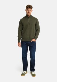 Oliven grøn sweater med høj krave og tre knapper, ribbede ærmekanter og kant, parret med mørke denim jeans og sneakers med grønne detaljer.