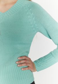 Világos teal színű, kötött pulóver V-nyakkal, bordás szegéllyel és textúrázott részletekkel a vállon. Hosszú, testhezálló ujjakkal rendelkezik.