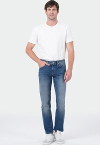 Homme debout portant un t-shirt blanc à manches courtes, un jean bleu et des baskets blanches, avec une main dans la poche, contre un fond clair.