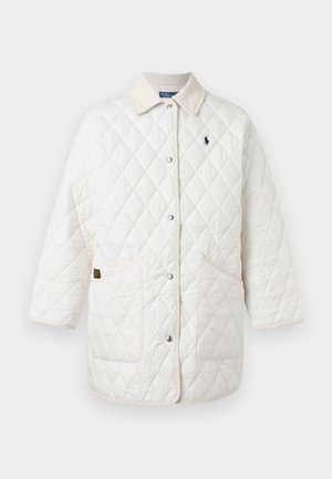 Person iført en hvid quiltet frakke, beige sweater, cremelange vidde bukser, hvide sneakers og en brun baseballkasket, stående med den ene hånd i lommen.