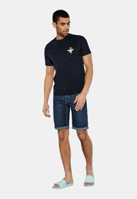 Maglietta di cotone blu navy con un logo geometrico, abbinata a pantaloni corti di denim arrotolati e sandali scivolati azzurri. Outfit estivo casual.