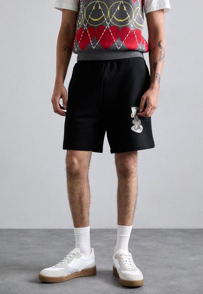 Black shorts with elastic waistband feature a metallic graphic on the leg. The outfit is paired with white sneakers and ribbed socks.