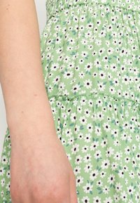 Vihreä kukkamekko, jossa on valkoisia kukkia ja mustia keskuksia. Mekon vyötärö on rypytetty ja hame on eteerinen. Kevyt kangas, jossa on pehmeä rakenne.