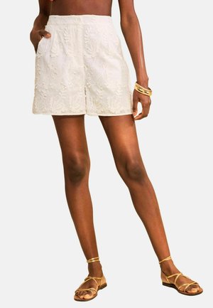 Frau trägt weiße, bestickte, hoch taillierte Shorts, goldene Armreifen und flache goldene Riemchensandalen, steht mit einer Hand in der Tasche.