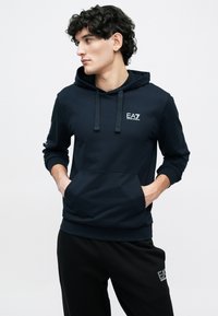 Tengerészkék kapucnis pulóver állítható húzózsinórokkal, kenguruzsebbel és mellkasi logóval. Fekete nadrággal párosítva, amelyen hasonló logó található az oldalon.