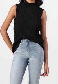 Schwarzes ärmelloses Oberteil mit hohem Kragen, ausgestattet mit vertikalen Falten. Kombiniert mit hellblauen, verwaschenen Skinny-Jeans. Geschmeidige Textur und lockerer Schnitt.