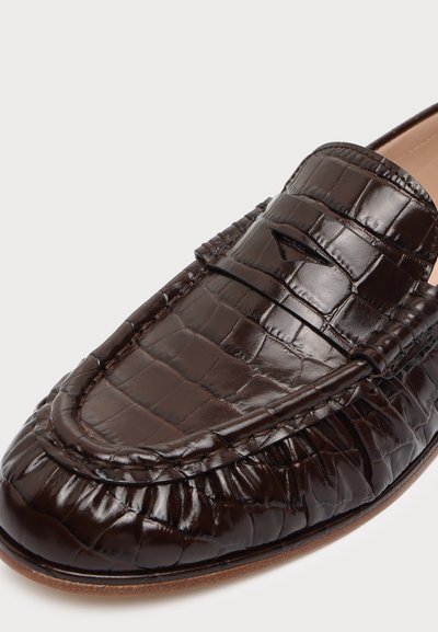 Gros plan sur un mocassin marron foncé en cuir brillant effet crocodile, avec un détail classique de fente à penny sur le dessus.