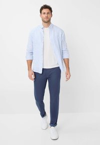 Camicia a righe blu chiaro e bianca con bottoni sopra una t-shirt bianca, abbinata a pantaloni blu navy e sneaker bianche, in piedi davanti a uno sfondo semplice.