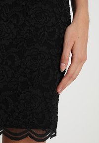 Main reposant à côté d'une jupe en dentelle noire avec des motifs floraux et un ourlet festonné sur un fond clair.