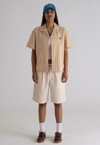Short-sleeved, button-up shirt with orange and white stripes, paired with beige shorts. A blue cap with logo, brown shoes, and white socks.