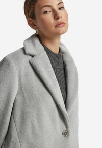 Grau melierter Wollmischmantel mit Reverskragen, einreihigem Knopfverschluss und weicher Textur, über einem gerippten grauen Pullover getragen.