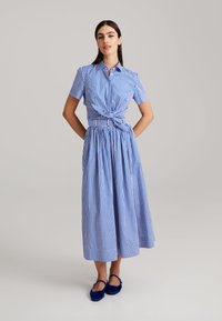 Blau-weißes Karokleid mit geknoteter Vorderseite, kurzen Ärmeln und einem voluminösen, Faltenrock. Getragen mit dunkelblauen Schuhen.