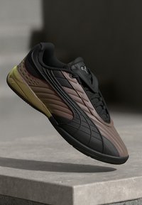 Tenis Puma maro și negru cu detalii galbene pe talpă, în aer deasupra unei suprafețe de piatră, pe un fundal întunecat neclar.