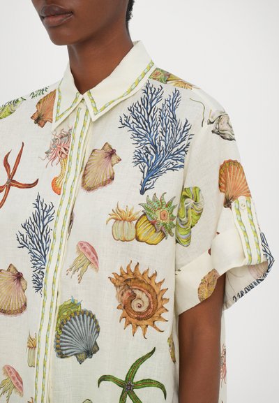 Camicia bianca in lino con illustrazioni marine colorate, tra cui conchiglie e coralli, e un dettaglio a righe in contrasto sul colletto.