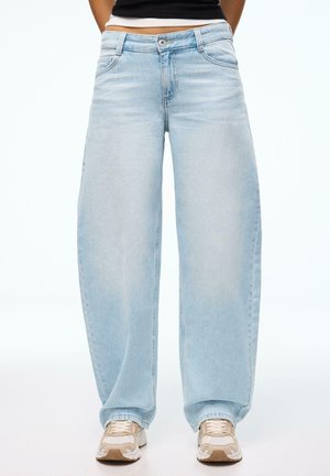 Person wearing light blue wide-leg jeans, black cropped top, and beige and white sneakers standing against a white background.