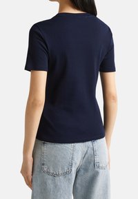 Marinblå bomullströja med korta ärmar, rund halsringning och en tight passform, sedd bakifrån mot ljusa jeans.