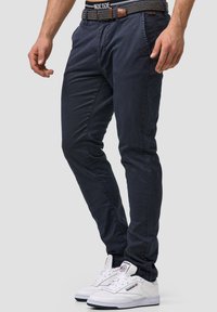 Navy blue stretch cotton chinos with a fitted design, featuring side pockets, and paired with white sneakers. Woven black belt with brown accents.
