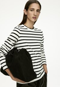Schwarze Wildleder-Bucket-Bag mit Kordelzugverschluss, gehalten von einer Person in einem schwarz-weißen gestreiften Langarmshirt.