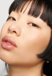 Nærbilde av et ansikt med jevn, lys hud og fyldige lepper. Sort rett hår med rett pannelugg. Brune øyne. Subtil glans på huden.