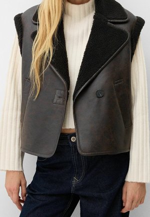 Femme portant un gilet en peau retournée en cuir marron foncé sur un pull crème côtelé et un jean bleu foncé, debout avec les mains détendues le long du corps.