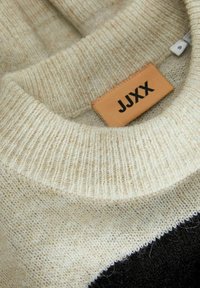 Strickpullover mit einem gerippten Rundhalskragen, beigem Material mit weicher Textur und einem Lederetikett, das "JJXX" zeigt. Akzentuiert mit Schwarz.