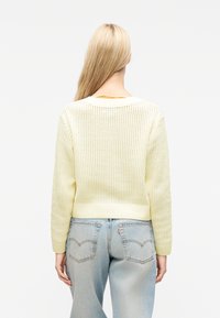 Ξανθιά γυναίκα με πλάτη στραμμένη, φοράει ανοιχτό κίτρινο πλεκτό πουλόβερ και ξεβάμμενο μπλε τζιν με ορατές πίσω τσέπες.