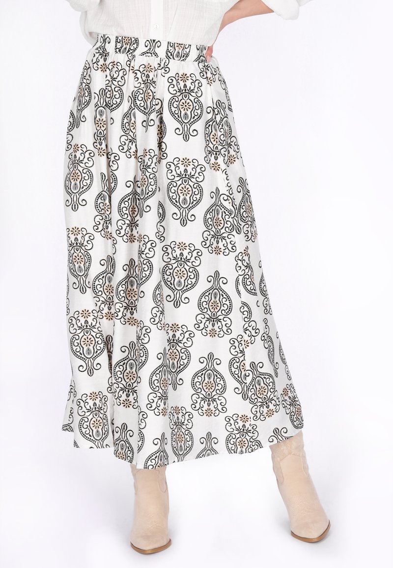 Weißer Midirock mit schwarzen und beigefarbenen, filigranen Blumen- und Wirbelmustern, kombiniert mit beigen Stiefeletten und einem weißen Oberteil.