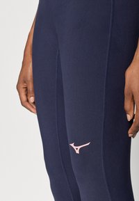 Tumši zilie sporta leggins aizstāti no elastīgas auduma ar rozā logotipu apakšējā kājā un plakano šuvju šūšanu komfortam.