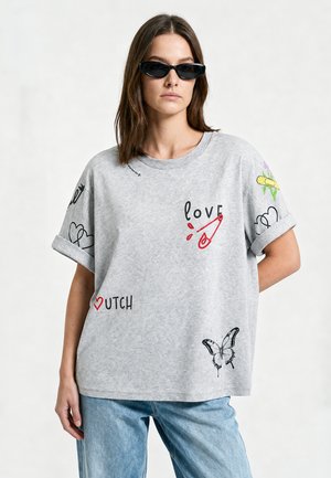 Femme portant des lunettes de soleil noires, un t-shirt gris avec un papillon, une épingle de sécurité, un cœur et les mots « amour » et « néerlandais », associée à un jean bleu.